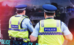 A New Zealand Police officer stands beside a patrol car on a residential street.