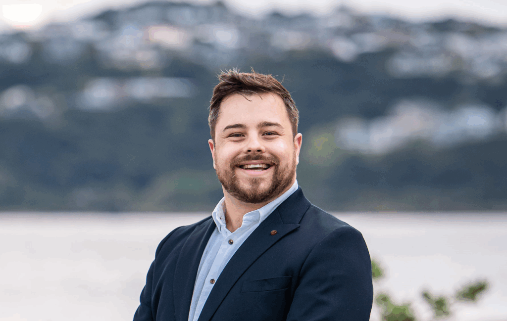 Josh Trlin smiling in a suit jacket with a blurred coastal background.