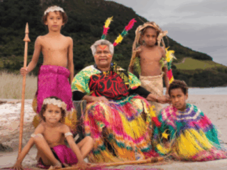 A Ni-Vanuatu elder in traditional dress sits on the beach with five children in colourful cultural attire, against a backdrop of coastal hills.