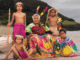 A Ni-Vanuatu elder in traditional dress sits on the beach with five children in colourful cultural attire, against a backdrop of coastal hills.