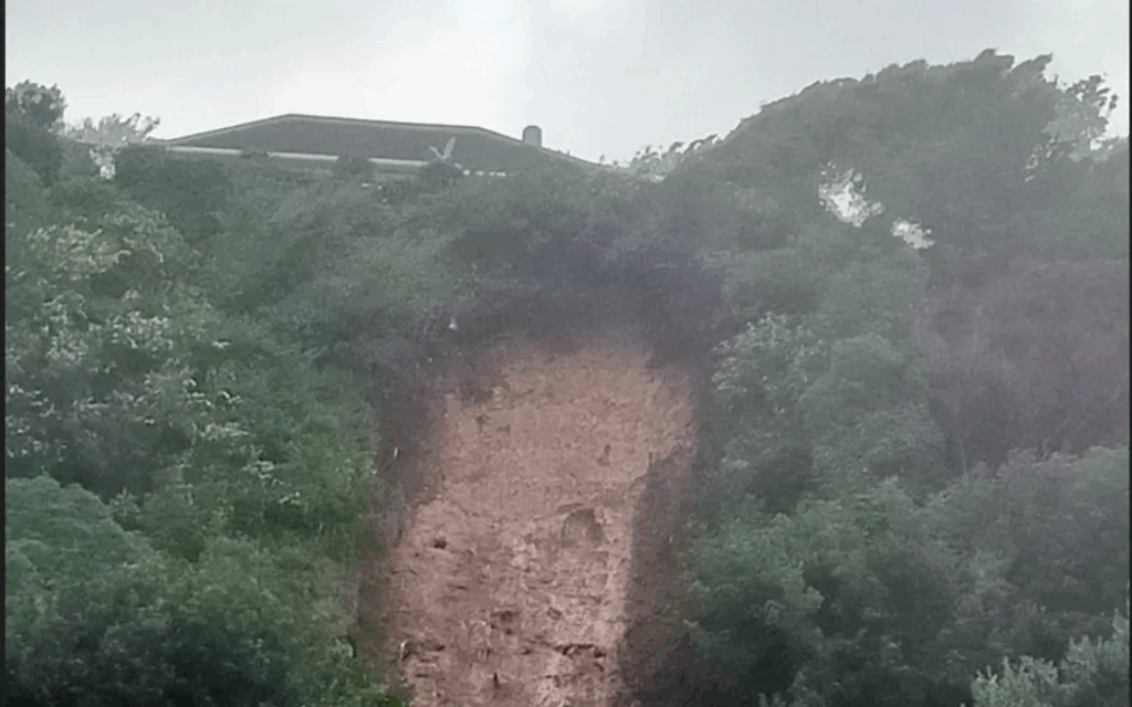 Muddy landslip debris piled against the rear walls of suburban houses in Porirua, with emergency cones and tape visible nearby.
