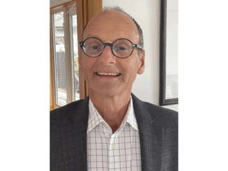 Grenville Gaskell wearing glasses and a checked shirt with a grey blazer, smiling indoors near a window.