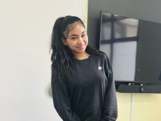 Teenage girl in black Nike sweatshirt, smiling and standing indoors.
