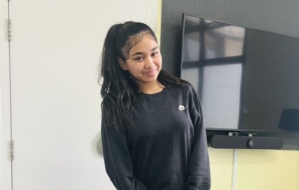 Teenage girl in black Nike sweatshirt, smiling and standing indoors.