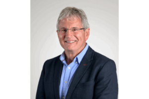 Ross Leggett wearing glasses, a blue shirt, and a dark blazer, smiling against a plain light background.