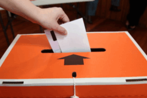 A hand places a voting paper into an orange and white Elections NZ ballot box during local elections.