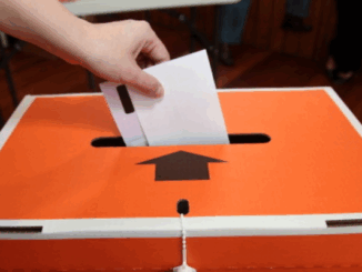 A hand places a voting paper into an orange and white Elections NZ ballot box during local elections.