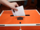 A hand places a voting paper into an orange and white Elections NZ ballot box during local elections.
