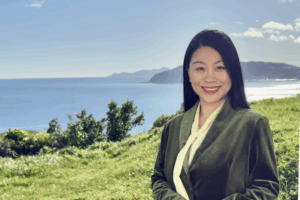 Yan Zang standing in front of a coastal landscape on a sunny day, smiling and wearing a dark green jacket with a cream blouse.