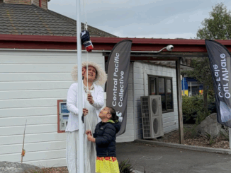 An adult in traditional Cook Islands attire and a child raise the Cook Islands flag outside a building with Central Pacific Collective banners.