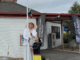 An adult in traditional Cook Islands attire and a child raise the Cook Islands flag outside a building with Central Pacific Collective banners.