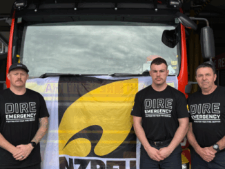 Three firefighters stand in front of a fire engine wearing "Dire Emergency" union shirts, protesting for better resourcing and support from Fire and Emergency NZ.