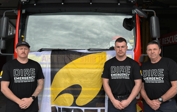 Three firefighters stand in front of a fire engine wearing "Dire Emergency" union shirts, protesting for better resourcing and support from Fire and Emergency NZ.