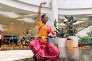 A woman dressed in vibrant traditional Indian attire performs a classical dance at North City Shopping Centre, surrounded by Diwali decorations and indoor plants.