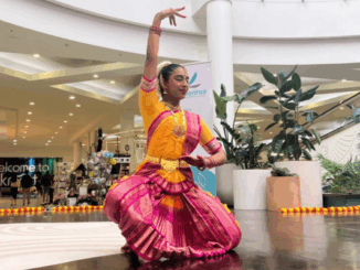 A woman dressed in vibrant traditional Indian attire performs a classical dance at North City Shopping Centre, surrounded by Diwali decorations and indoor plants.
