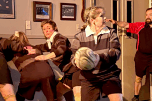 Four women dressed in rugby uniforms act out a scene in a clubroom setting, with one holding a rugby ball and others engaged in dramatic play, while a man in a referee shirt looks on from the background.