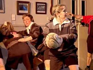 Four women dressed in rugby uniforms act out a scene in a clubroom setting, with one holding a rugby ball and others engaged in dramatic play, while a man in a referee shirt looks on from the background.