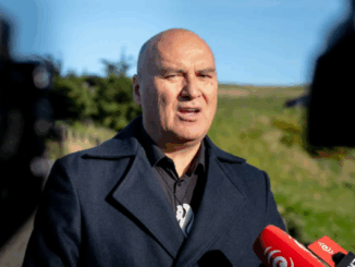 Helmut Modlik, Chief Executive of Ngāti Toa Rangatira, speaks outdoors at a media event. He is wearing a dark coat and standing in front of grassy hills, with red RNZ microphones visible in the foreground.