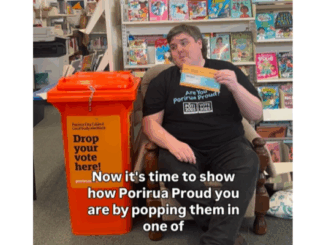 Porirua Deputy Electoral Officer Jack Marshall sitting beside a bright orange ballot drop box in a library, holding a voting envelope. Text on screen reads: “Now it's time to show how Porirua Proud you are by popping them in one of...”