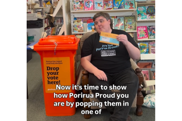 Porirua Deputy Electoral Officer Jack Marshall sitting beside a bright orange ballot drop box in a library, holding a voting envelope. Text on screen reads: “Now it's time to show how Porirua Proud you are by popping them in one of...”