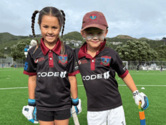 Two children wearing North City Cricket Club uniforms smile on a cricket field with trees and hills in the background