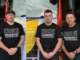 Three firefighters wearing “Dire Emergency” union shirts stand in front of a fire truck draped with an NZPFU banner, preparing for a strike to raise awareness about working conditions.