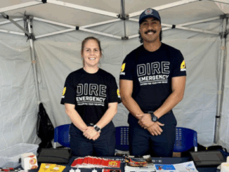 Two Porirua firefighters standing and smiling inside a white event tent, wearing “Dire Emergency – Fighting for Your Fire Service” union t-shirts. They are behind a table with fire safety pamphlets and promotional materials.