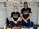 Two Porirua firefighters standing and smiling inside a white event tent, wearing “Dire Emergency – Fighting for Your Fire Service” union t-shirts. They are behind a table with fire safety pamphlets and promotional materials.