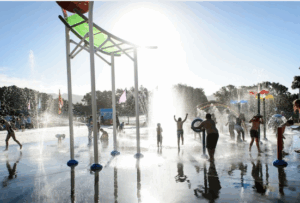 Kids playing among colourful water fountains and splash buckets at Aotea Lagoon’s water play area in Porirua on a bright summer day.