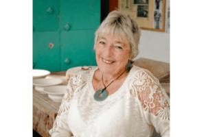 Anneke Borren smiling in her studio, seated beside shelves of pottery and tools.