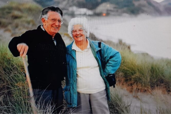 Elderly couple smiling at beach dunes; man holding walking stick, woman wearing turquoise jacket and glasses, coastal hills and sea in background.