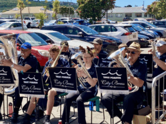 Members of Porirua City Brass and Porirua Youth Band perform together outside The Warehouse, seated with brass instruments as shoppers and parked cars fill the background.