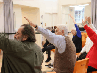 A group of older adults seated in chairs raise one arm during a gentle exercise class in a community hall.
