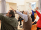 A group of older adults seated in chairs raise one arm during a gentle exercise class in a community hall.