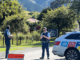 Two Police officers stand beside a marked patrol car at a road cordon on Paekākāriki Hill, with rural fencing, trees and bush-covered hills in the background.