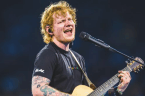 A male musician sings into a microphone while playing an acoustic guitar on a brightly lit stage.