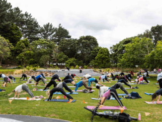 People take part in an outdoor BodyBalance class on the grass at Aotea Lagoon, holding yoga-style poses on mats.