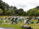 People take part in an outdoor BodyBalance class on the grass at Aotea Lagoon, holding yoga-style poses on mats.