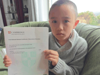 A young boy sits on a couch holding a Cambridge International Education certificate for mathematics.