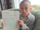 A young boy sits on a couch holding a Cambridge International Education certificate for mathematics.