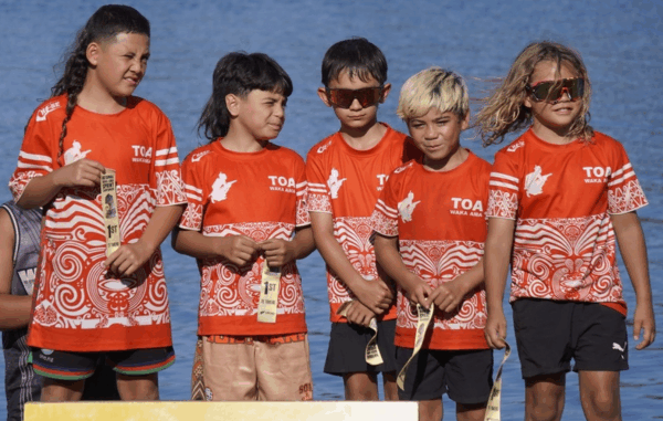A group of young TOA Waka Ama paddlers wearing red team shirts stand together on a podium holding medals, with water visible behind them.