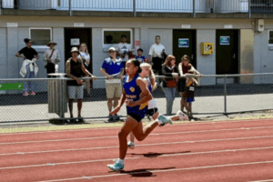 A Titahi Bay junior athlete runs on the track at Newtown Park Stadium, with spectators watching from behind the fence during a junior athletics meet.