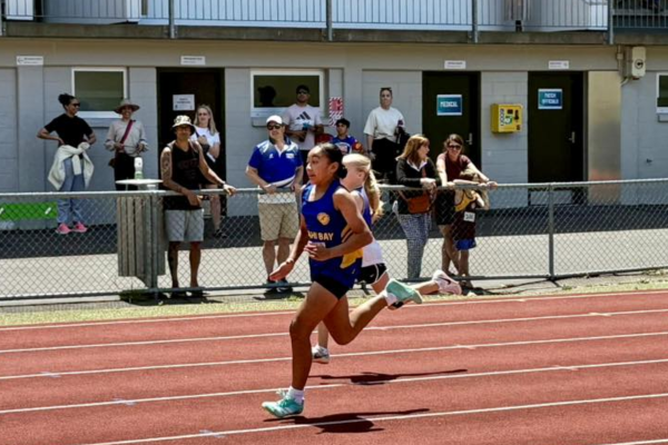 A Titahi Bay junior athlete runs on the track at Newtown Park Stadium, with spectators watching from behind the fence during a junior athletics meet.