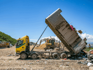 A yellow dump truck tips a large load of rubbish onto a landfill site while heavy machinery operates in the background under a clear blue sky.