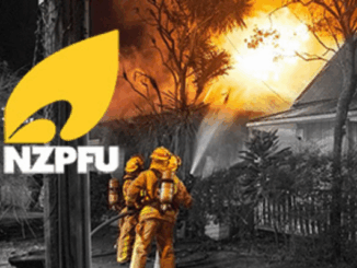 Two firefighters in yellow protective gear spray water on a house fire at night as flames and smoke rise behind trees, with the NZPFU logo displayed in the corner.