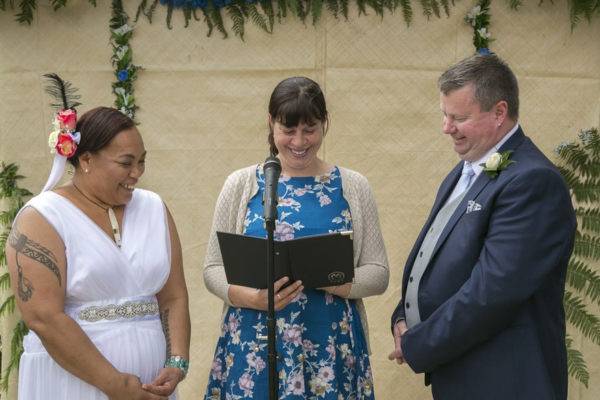 Wedding celebrant standing at a microphone reading from a folder during an outdoor ceremony while a smiling couple stands on either side.