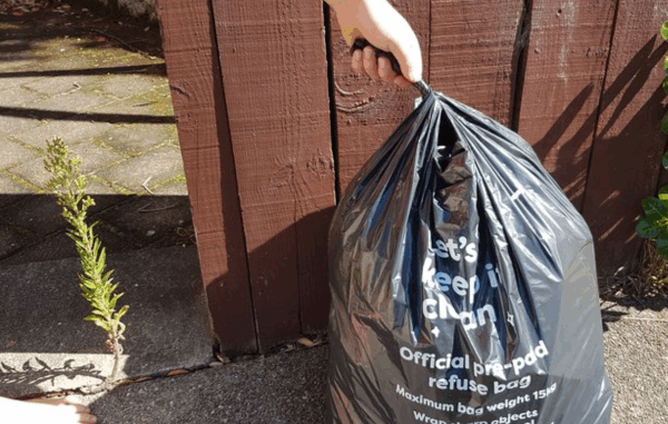 Hand holding a black official Porirua pre-paid rubbish bag beside a wooden fence at a residential kerbside.