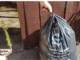 Hand holding a black official Porirua pre-paid rubbish bag beside a wooden fence at a residential kerbside.