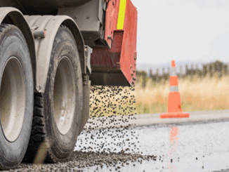 Truck spreads chipseal on wet road surface during highway maintenance works.