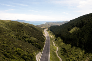 Aerial view of Transmission Gully motorway winding through green hills toward the Kāpiti Coast, with the sea visible in the distance.